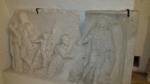 Mészkő relief; sírépítmény dísze. Töredékes, a bal oldali kétharmad rész a Rév-komáromi Dunamenti Múzeumban található. A hiányzó rész mindkét helyen egymást kiegészítő gipszmásolat.